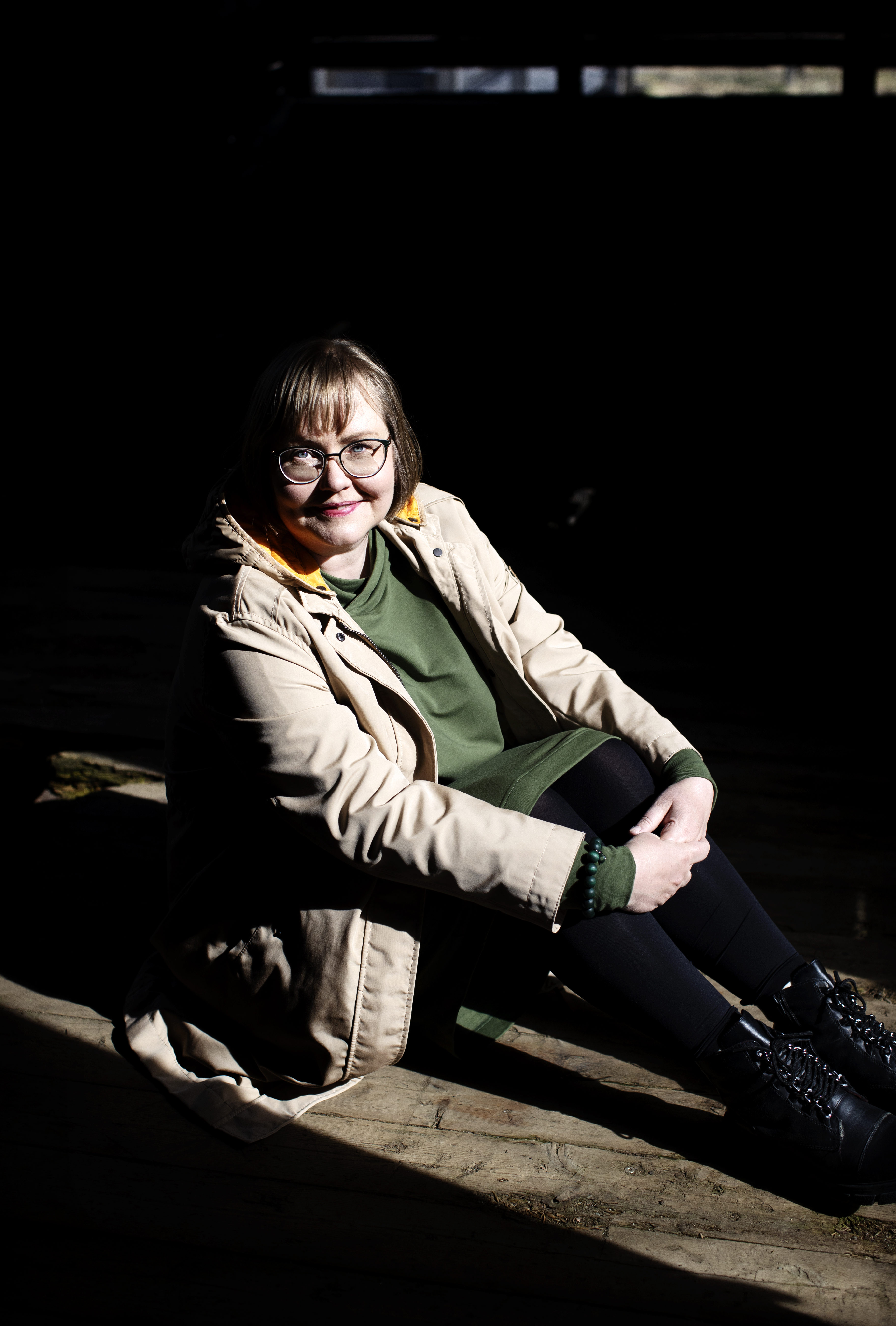 Silmälasipäinen Sonja Miettinen istuu valaistuna pimeässä ympäristössä muurin päällä ja hymyilee. Kuvan on ottanut Heidi Piiroinen.