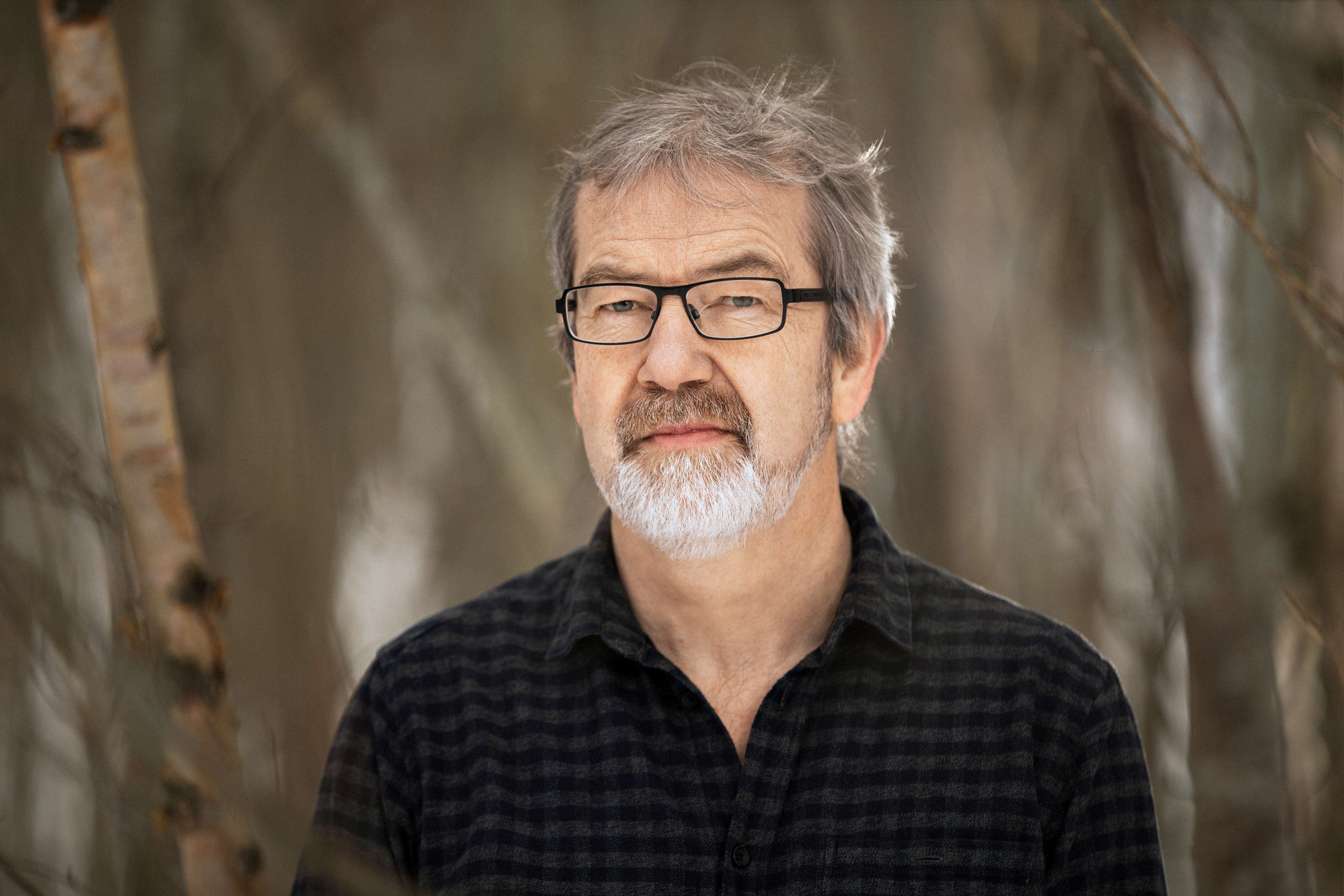 Kuvassa harmaapartainen ja harmaahiuksinen Jan Tossebro. Hänellä on musta paita ja mustat silmälasit. Taustalla koivun runkoja.